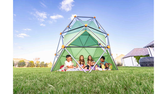 Multiple kids smiling under a climbing dome with a tent on a sunny day.